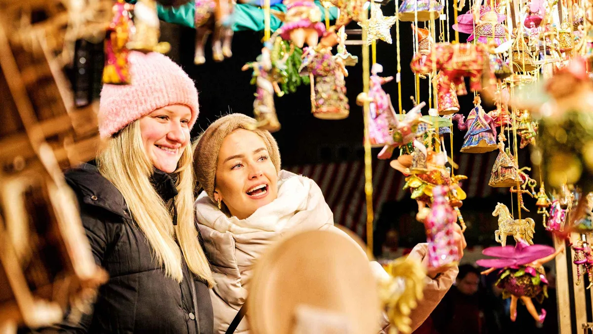 Two women smiling at a stall hung with glass ornaments and wooden toys